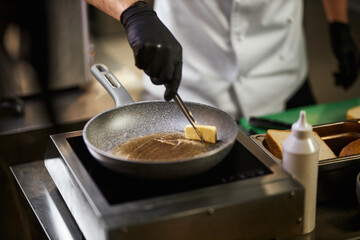Crope view of chef's hands in black gloves melting piece of butter in frying pan. Male in chef...