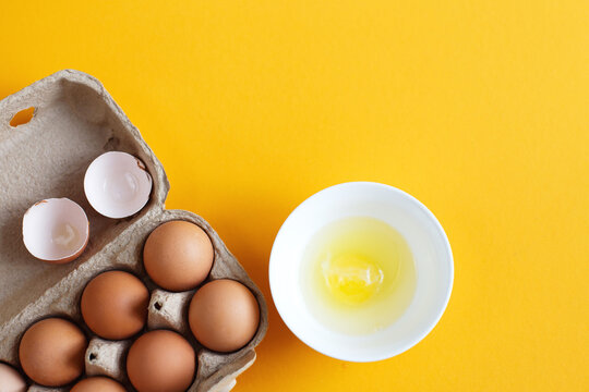 raw eggs in a package next to white bowl with one broken egg in it on a yellow background - Powered by Adobe