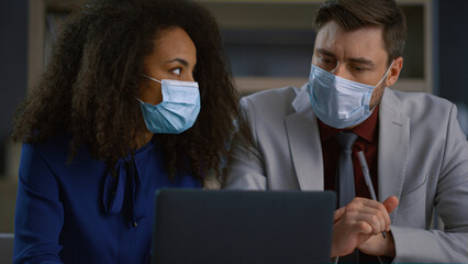 Diverse businesspeople wear mask analyzing data on laptop online in covid office