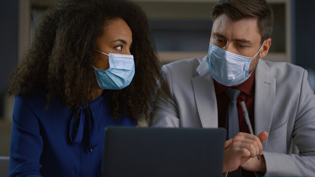 Diverse businesspeople wear mask analyzing data on laptop online in covid office