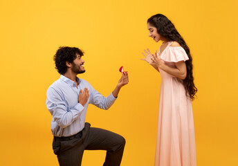 Loving indian man standing on one knee and offering engagement ring to his beloved woman on yellow...