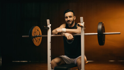 Obraz na płótnie Canvas Smiling sportsman looking at camera near barbell on stand on dark background