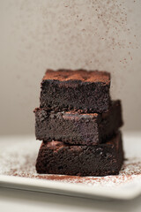 Stack of homemade chocolate brownies under falling cocoa powder on a saucer, gray table and background with place for text. Vertical photo with shallow depth of fields.