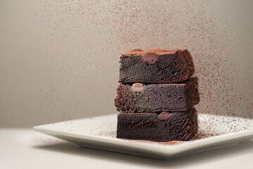 Stack of homemade chocolate brownies under falling cocoa powder on a saucer, gray table and background with place for text. Horizontal photo with shallow depth of fields.
