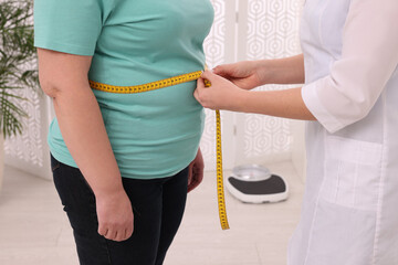 Nutritionist measuring overweight woman's waist with tape in clinic, closeup