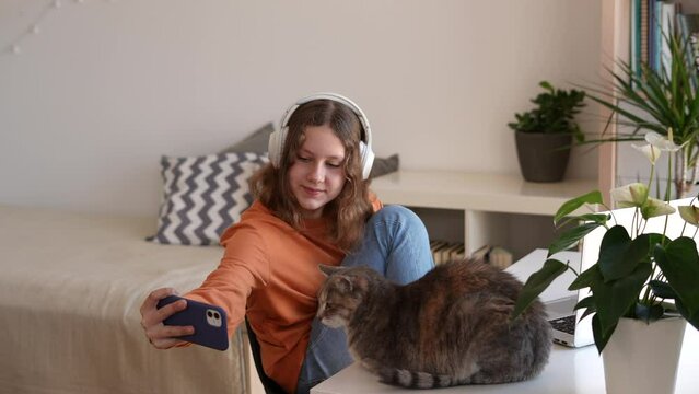 Adorable teenage girl with wireless headphones sitting at the table taking selfie with her pet cat to make a funny photo. 