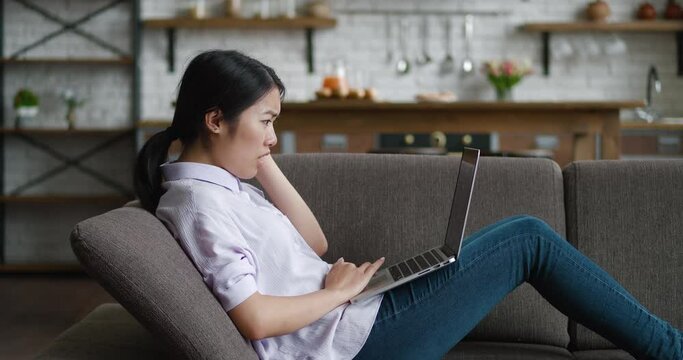 Shocked asian business woman looks at laptop with fear of terrible news, while resting on sofa at living room. Portrait of scared female student, reaction on bad news, sitting indoors at home