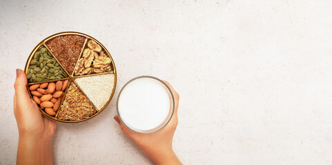 Women's hands hold a glass of vegetable milk and a plate divided into sectors with various seeds and nuts. Concept: alternative, vegetable milk. Healthy eating.