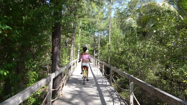 Following shot of cute girl riding a bicycle on the rural wooden bridge in beautiful tropic nature 