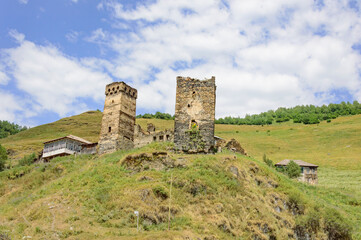 Settlement with two medieval svan towers on the road to Ushguli medieval fortified village in Svanetia, Georgia. Green mountain slopes, ols yellow and grey stone walls, blue sky with clouds