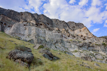 Vardzia ruined rock-hewn city in Georgia. Grey and yellow rock cliff with numerous caves, green nountain slopes, blue sky with clouds