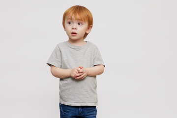 Cute red-haired and red-headed boy in grey T-shirt on grey or white background folding his hands and looking a bit scared to the left side. Standing isolated looking adorable. Copy space. 
