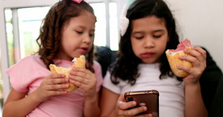 Two little girls hypnotized by cellphone while eating sandwich snack