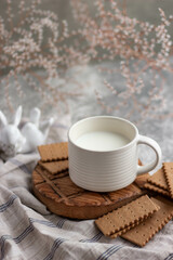 Obraz na płótnie Canvas A white ceramic mug with milk, biscuits and a white ceramic bunny surrounded by small white flowers. A gentle breakfast in a light key