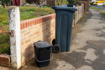 A black wheelie bin and waste food caddy on a pavement outside the front garden wall.