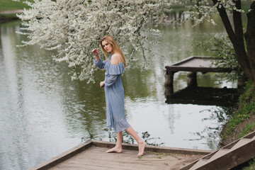 Young pretty caucasian blonde woman with freckles wearing natural makeup in light blue dress near the beautiful blooming spring tree by the lake. Youth, freshness, beauty, happiness, emotions concept.