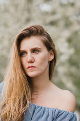 Fototapeta premium Young pretty caucasian blonde woman with freckles wearing natural makeup in light blue dress near the beautiful blooming spring tree by the lake. Youth, freshness, beauty, happiness, emotions concept.
