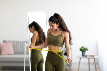 Fit Indian woman in sportswear measuring waist with tape near mirror, satisfied with slimming...