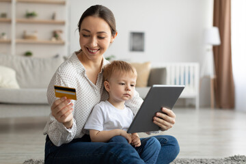 Young mother with child boy using digital tablet and credit card for internet purchases, sitting on floor in living room