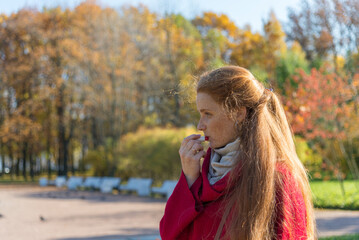 Beautiful young woman with long red hair and freckles on her face paints her lips with lipstick in a sunny autumn park