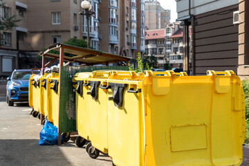 Rows of many big plastic yellow dumpster cans full of black plastic trash litter bags near residential building at city downtown or suburban area. Non-recyclable sorting garbage collecting