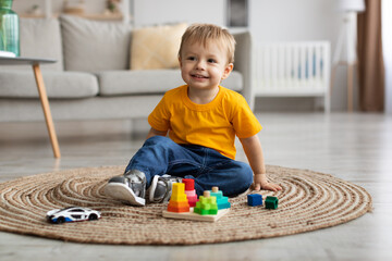 Cute toddler boy playing with car and educational wooden toy at home, sitting on floor carpet in...