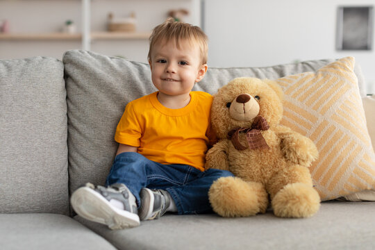 Cute little toddler boy sitting on sofa near teddy bear and looking at camera, adorable child playing with toy