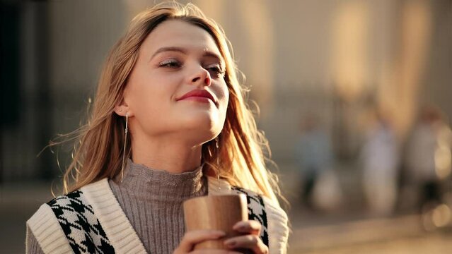 Girl taking a sip of coffee or tea from wooden eco cup. Happy Beautiful Blonde woman enjoying cup of coffee or tea on warm summer evening sunset in big city