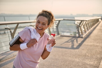 Happy delightful mixed race female athlete runner holding a bottle with water and a white towel on...