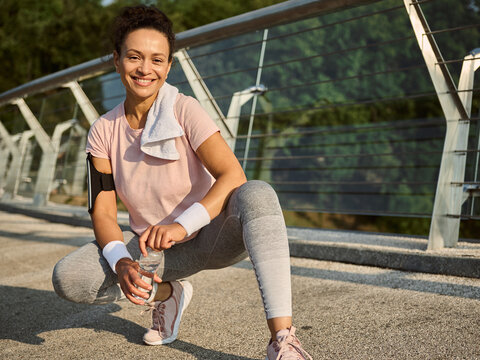 Portrait of female athlete in sportswear and white terry wristbands holding a bottle of water sitting on squat pose on a treadmill, smiles looking at camera relaxing after morning jog along the bridge