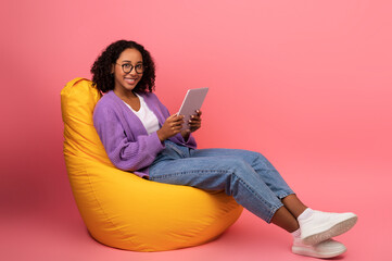 Full length of joyful young black woman with digital tablet sitting on bean bag chair, working...