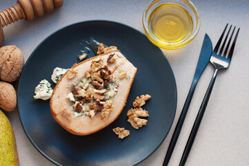 Pear baked with dorblu cheese, honey and walnuts on a black plate on a gray background. Backed pear with fork and knife. Top view