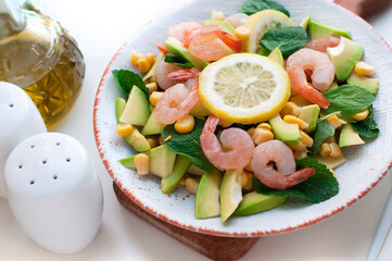 Salad with shrimps, avocado, corn and lemon with olive oil on a wooden board on white background. Salad with shrimps and avocado on white plate with fork and knife