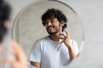 Happy indian man looking in mirror spraying on cologne in bathroom interior, doing morning skincare...