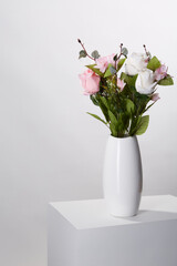 Subject shot of graceful bouquet of tender pink and white roses in a white ceramic vase. The vase with bunch of flowers is located on the white table on the white background. 