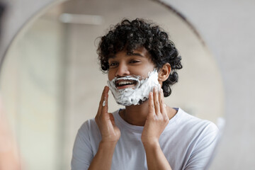 Shaving routine concept. Indian man applying shave foam on face making morning hygiene and looking...