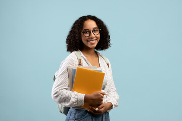 Academic education. Pretty black female student with backpack and notebooks smiling at camera over blue background
