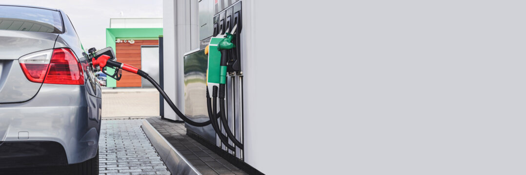A gun in a car tank at a gas station. Against the background of a column with fuel hoses of binzin, diesel, eco hydrogen. Long banner.