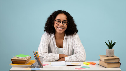 Happy young black female student sitting at desk with books and notebooks, smiling at camera on blue background