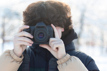 Fototapeta premium A teenage boy with curly long hair takes a picture with a digital SLR camera. Winter photo shoot outdoors