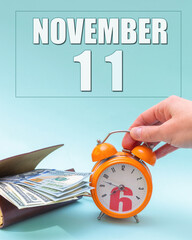 November 11th. Hand holding an orange alarm clock, a wallet with cash and a calendar date. Day 11 of month.