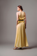 Young ginger lady in yellow evening dress with pleating skirt, high halter neckline and black belt on thin waist, rear view. Female bright look on grey studio background, glamour style.