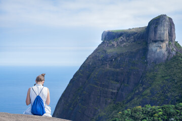 A woman traveler with a backpack sits against the background of an unusual rock, sky and ocean