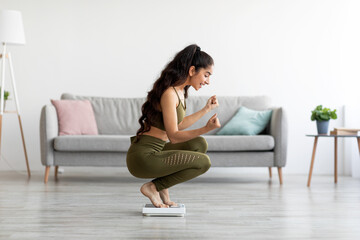 Triumphant millennial Indian woman sitting on scales, making YES gesture, excited about her weight...