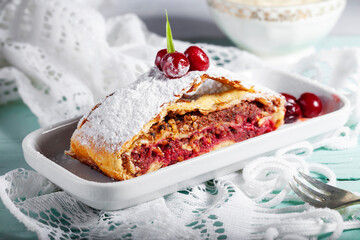 
A piece of freshly baked homemade strudel with cherries and powdered sugar, on a rectangular white porcelain plate on a light background