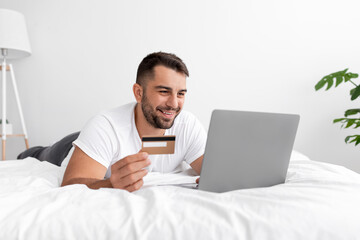 Cheerful relaxed young caucasian muscle man lies on white bed, looks at computer, holds credit card