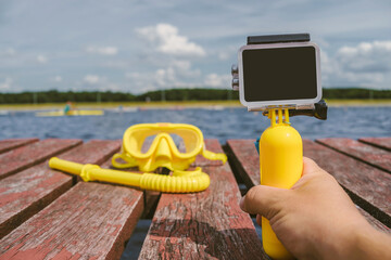 Close-up, action camera mockup with a yellow float in a man's hand. Against the background of a waterproof mask for snorkeling, diving with a tube on a wooden pier with water.