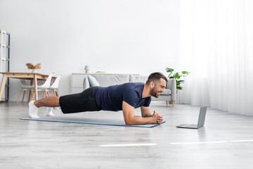 Obraz premium Smiling young caucasian guy doing plank on mat on floor and look at laptop in living room interior, profile