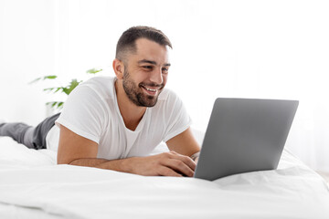 Happy smiling young european muscular man lies on white bed and chatting on computer, watch funny video
