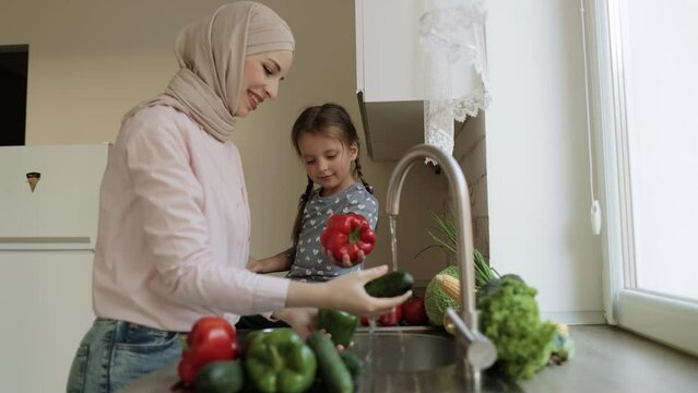 Happy Loving Family Preparing Food Together. Smiling Arabian Mom In Hijab And Child Daughter Girl Washing Vegetables Pepper And Cucumber And Having Fun In The Kitchen. Little Helper. Healthy Food